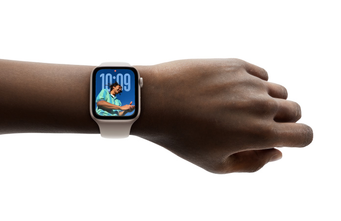 고무 밴드를 장착한 로즈 골드 마감의 Apple Watch가 손목에 착용된 이미지, 새로운 Liquid Glass 디자인의 시계 앱을 보여주는 모습
