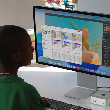 員工坐著使用 iMac 和 Mac Mini 上名為「Q4 Sales Tracker」的 Excel 試算表。Excel 試算表視窗後方有一個 Pixelmator Pro 視窗和 macOS Tahoe 背景圖片。